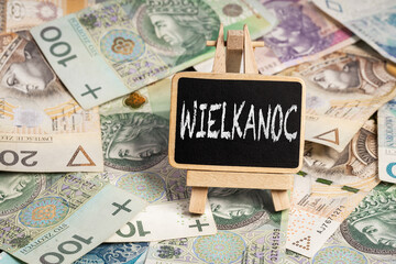 Black writing board on a wooden frame with the inscription "wielkanoc", Polish zloty PLN banknotes scattered in the background (selective focus) translation: easter
