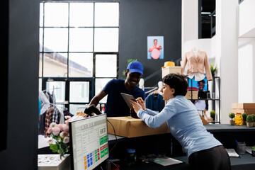 African american courier discussing shipping details with employee, checking online order in on tablet computer. Store worker standing at counter desk, preparing packages for delivery in boutique