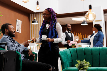 Hotel waitress holding cup of freshly brewed coffee taking cash payment from guest while serving...