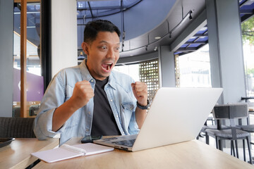 Fototapeta premium Asian man happy and enthusiastic with blue shirt using laptop and mobile phone in coffee shop, online shopping business