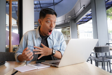 Asian man shocked face, wow, surprised with blue shirt using laptop and mobile phone in coffee...