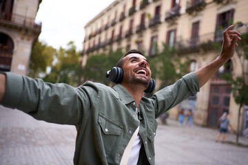 Young adult Caucasian man with wireless headphones on stands on European Barcelona city street with...