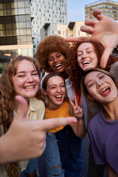 Happy vertical selfie of an multiracial group of female friends making frame gesture with fingers outdoors in the city. Young women looking at camera smiling.