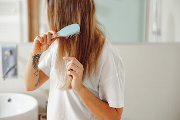 Woman standing in front of mirror at bathroom and combing her hair with brush after shower 