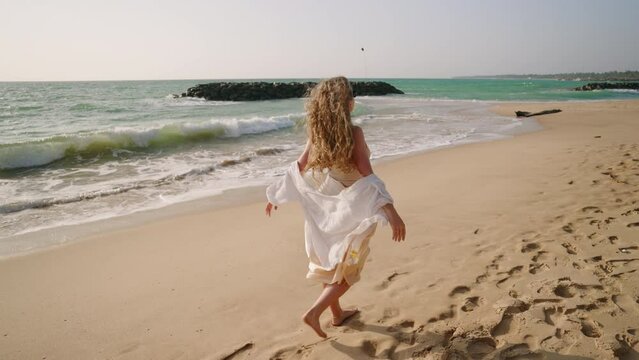 Young pretty woman going barefoot, catching wind, leaving footprints on sand at beach. Curly girl walking at seaside, foamy water waves. Caucasian model posing, hair blowing in wind at the coast.