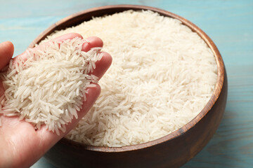 Woman holding raw basmati rice over bowl at light blue wooden table, closeup