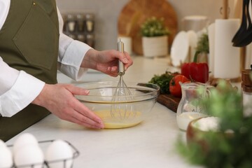 Woman whisking eggs in bowl at light table indoors, closeup