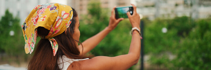 Cute tanned woman with long brown hair wearing white top and yellow bandana standing on bridge taking photo of modern city, Panorama