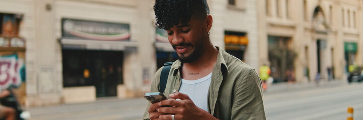 Young smiling man with beard dressed in an olive color shirt uses phone map app on the old city background, Panorama