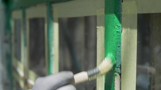 Hand wearing glove is painting wooden window with green paint. Applied with brush. Patch of old wooden surface, showing through cracked and flaking paint. Handiwork, restore woodwork outdoor