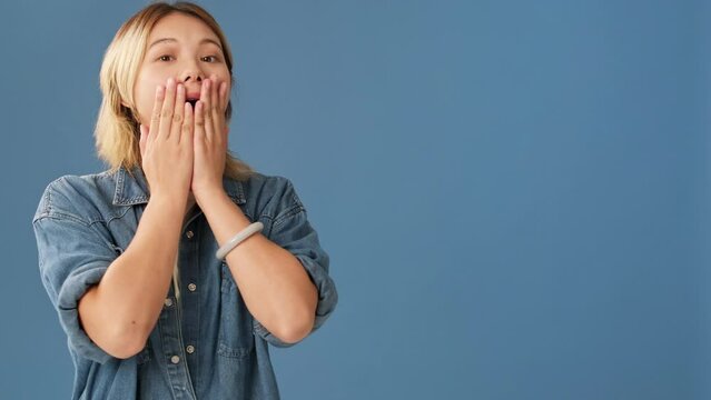 Surprised woman looks to side covering her mouth with her palms isolated on blue background