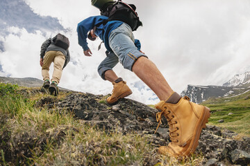 Two young hikers with backpacks walks in mountains. Close up photo of tourist legs going uphill - obrazy, fototapety, plakaty