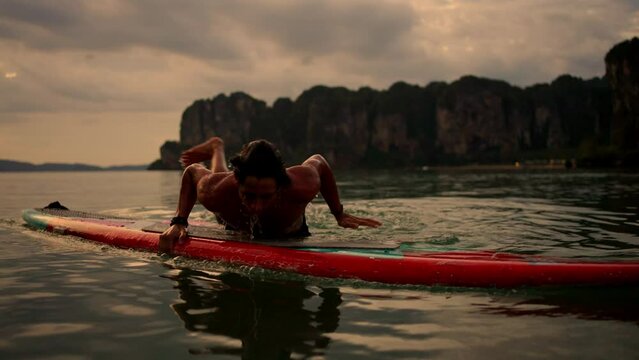 4K Young Asian man swimming and rowing paddle board in the ocean at tropical island at sunset. People enjoy outdoor active lifestyle water sport surfing and paddle boarding on summer beach vacation.