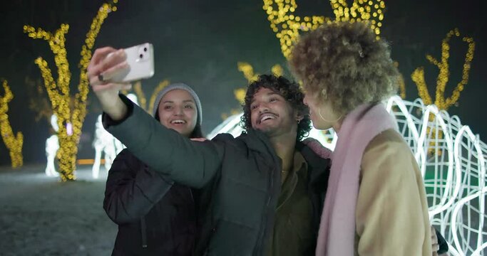 Excited happy friends taking selfie on smartphone on garlands and lights background - New Year holidays Christmas vacation
