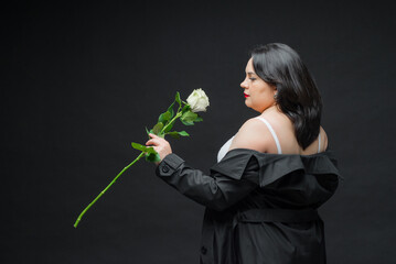 A beautiful girl in a black cloak on a dark background holds a white rose in her hand. The image...