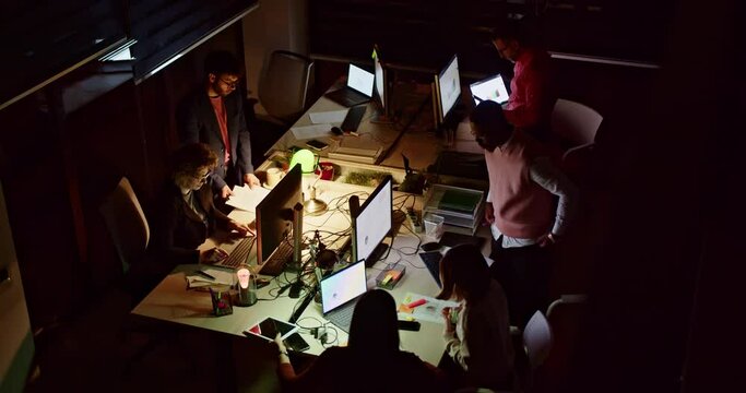 A focused group of business professionals working at their desks in a dark office illuminated by computer screens, collaborating efficiently beyond regular work hours.