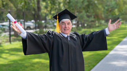 An elderly gray-haired man in a graduate robe spread his arms to the sides and holds a diploma...