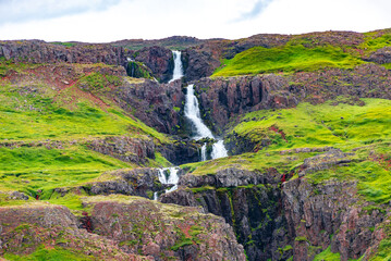 Cascading waterfall in East Iceland from fjord top, with dramatic sky and green meadow hills....