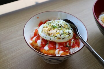 A bowl filled with rice, chicken katdu and eggs on the top. Served on wooden table. Sauce and mayonaise on the top.