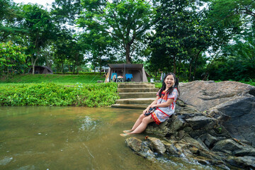 camping tent by asian child camper happy smile or kid girl sitting relax on rocks and water stream to people travel in nature trees garden forest on summer holiday vacation at Ratchaburi in Thailand