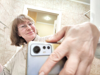 Fototapeta na wymiar Mature woman captures her reflection while taking a selfie. The reflection of a middle aged girl with a phone in the mirror. Partial focus