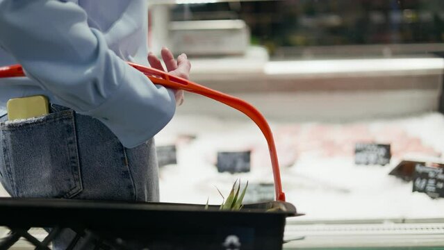Unrecognizable person in jeans holding shopping basket while standing by open refrigerator with price tags. Elite store consumer keeping cell phone in back pocket of pants while considering purchase.