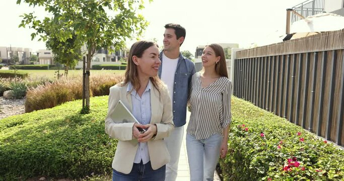 Hispanic married couple follow female realtor walking along bushes enjoy garden and well-groomed view of potential accommodation, listening agent speech, entering inside inner yard of private villa