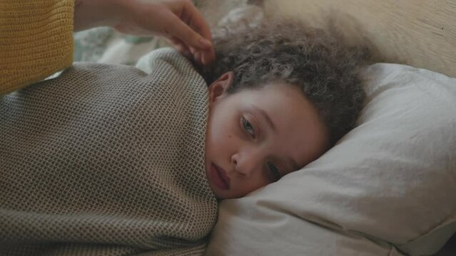 A sick baby girl lying in bed with a fever, mama is caring for a sick daughter. Face of hispanic curly hair ill female child kid taking a nap, mother touching a head, giving support 