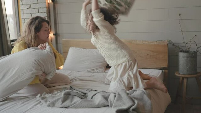 Happy active family mom and little child girl enjoy funny pillow fight on bed, smiling mother with small cute kid daughter having fun together playing laughing in bedroom together in the morning