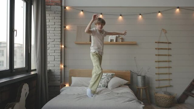 Little carefree boy jumps on bed in modern light bedroom, lovely kid pretend, imagining like fly in air, have fun alone indoor. Weekend leisure, hyperactive child, free time activity at home concept.