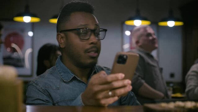 African American man rejoices at the soccer team victory in championship, checks bookmaker ratings online using phone. Happy sport fan wins bet on football match sitting in bar or sports pub at night.