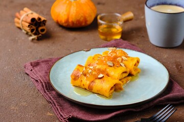 Thin pumpkin pancakes rolled into a roll and topped with honey and jam on a gray ceramic plate on a brown concrete background. Pancake recipes.