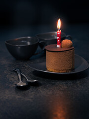 Chocolate cake with burning candle, two coffee cups and spoons on dark table against black background