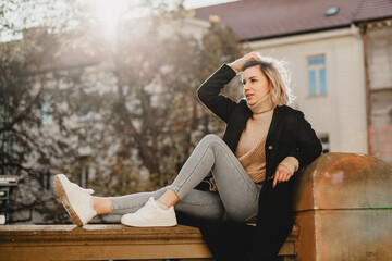 Stylish Woman in Black Jacket and White Shoes Sitting on Ledge. Urban Fashion Portrait with Trendy...