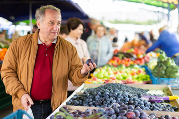 Obraz premium Positive mature male holding plum while choosing fruits and vegetables at market
