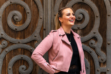 woman wearing a pink jacket and black pants stands in front of a wooden door