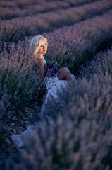 Blonde woman poses in lavender field at sunset. Happy woman in white dress holds lavender bouquet. Aromatherapy concept, lavender oil, photo session in lavender