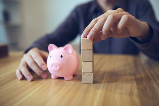 Person building a tower from wooden blocks beside a piggy bank, symbolizing financial planning and saving