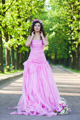 Fototapeta premium Charming female model with bright makeup, curly hair and pink flower wreath on green summer foliage background. Healthy woman in pink dress portrait