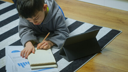 Cute kid lying on the floor studying with tablet computer writing in notebook. Child do homework at home