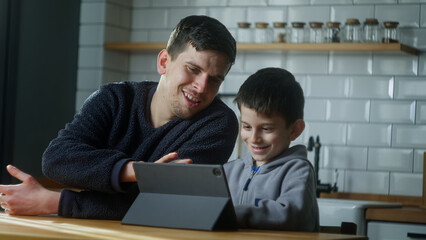 Young dad and son having fun using digital tablet sitting in the kitchen. Cute kid learning technology look at pad screen at home.	

