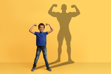 A cheerful boy in a blue shirt flexes his muscles with a big smile, casting a strong superhero silhouette