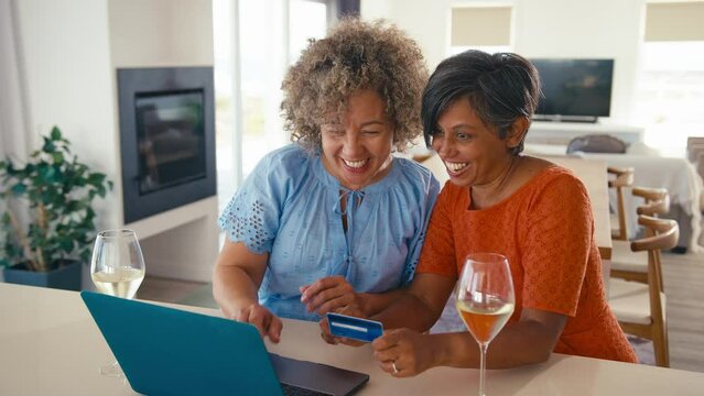 Two excited mature female friends with laptop at home using credit card to book vacation tickets or shop online celebrating drinking wine - shot in slow motion