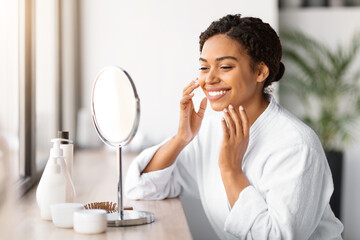 Happy young black woman applying facial moisturizer while looking in makeup mirror