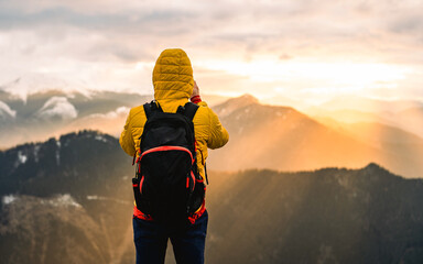 Hiker meets the beautiful sunset. Traveler hiking in mountain forest wearing cold weather accessories, wind jacket and backpack for camping outdoo - obrazy, fototapety, plakaty