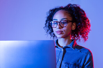 Focused black woman with glasses working on laptop with blue lighting