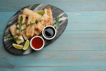 Tasty fried spring rolls, arugula, lime and sauces on light blue wooden table, top view. Space for...