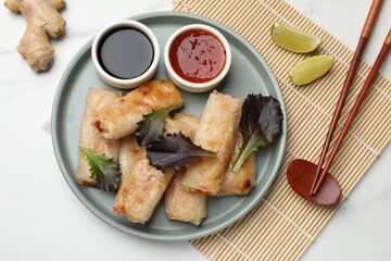 Tasty fried spring rolls, lettuce, sauces and other products on white marble table, flat lay