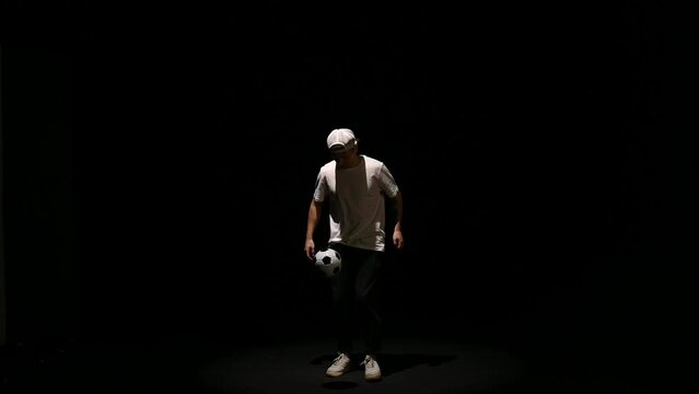 A young man throws a soccer ball on a black background. Football player shows tricks with the ball. Freestyler hits a black and white ball on a dark background. High quality FullHD footage