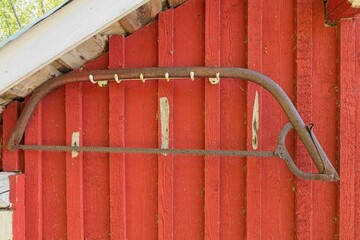 Closeup of old rusty bow saw (swede saw, finn saw or bucksaw) hanging on wooden wall of traditionally red painted building.